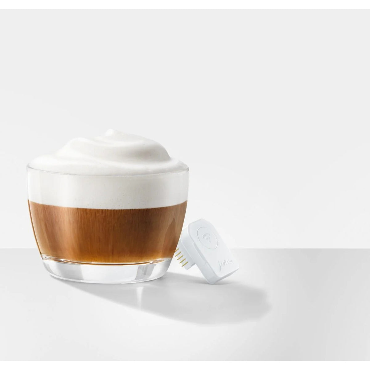 JURA WiFi Connect V2 (24323) bílý modul položený vedle sklenice s cappuccinem, nahoře pěna