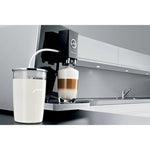 JURA Glass milk container (72570) next to coffee machine, transparent glass container with milk and silver lid on kitchen counter