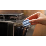 Hand inserting a light-blue descaling tablet into a coffee machine reservoir; shiny black appliance visible