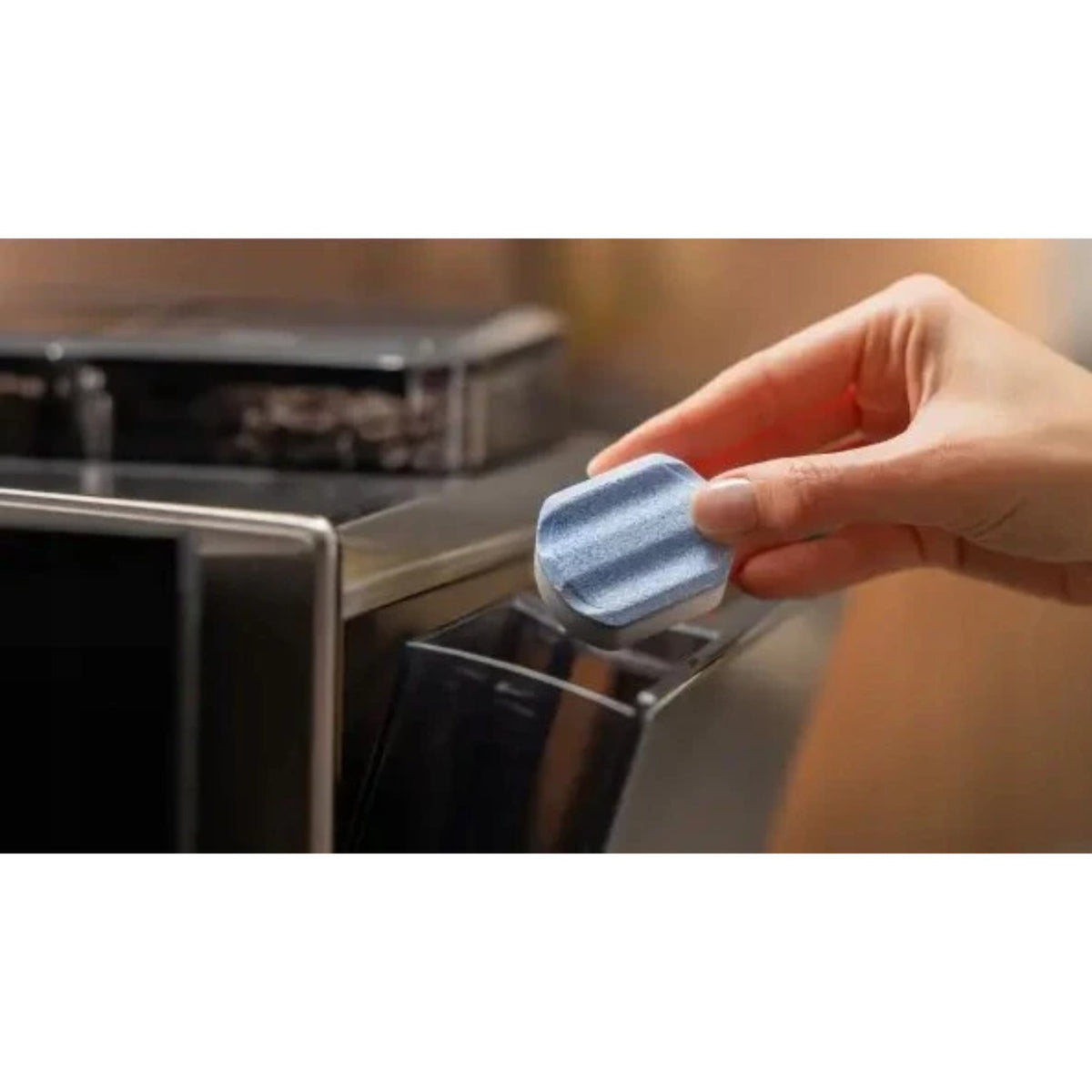 Hand inserting a light-blue descaling tablet into a coffee machine reservoir; shiny black appliance visible