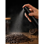 Small coffee bean sprayer 10ml black, being sprayed by hand over a pile of glossy coffee beans in a fine mist